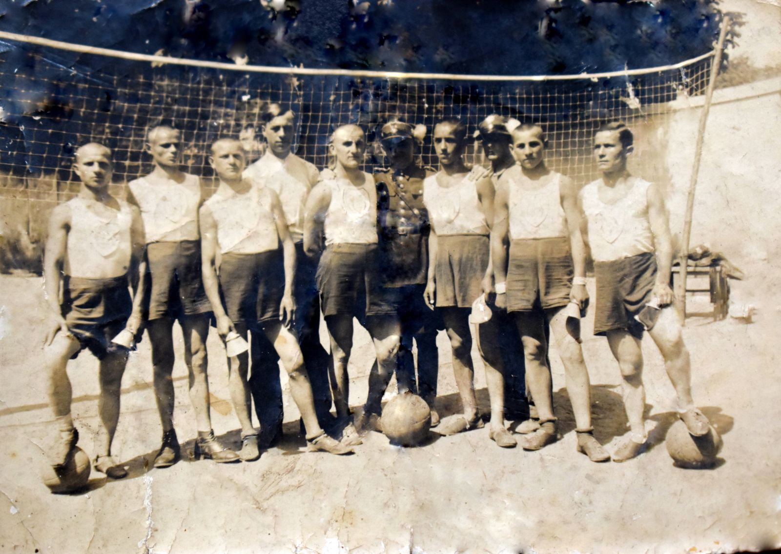 Zdjecie archiwaln na tle bramki do piłki nożnej stoi  siedmiu mężczyzn  ubrani w szirty i bluzeczki na ramiaczkach w środku grupy dwóch mężczyzn ubranych w mundur wojskowy