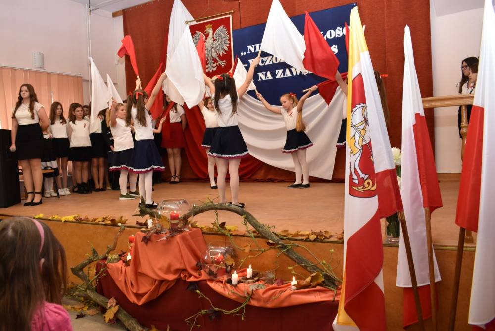Sala w Domu Kultury, scena na niej dziewczęta i chłopcy ustawieni w pólkolu,w środku dziewczynki w białych bluzkach i granatowych spódniczkach stoja w kole, trzymają do góry uniesione flagi narodowe